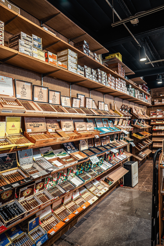 Wooden display shelves stocked with cigar boxes and individual cigar packs in a cigar shop interior.