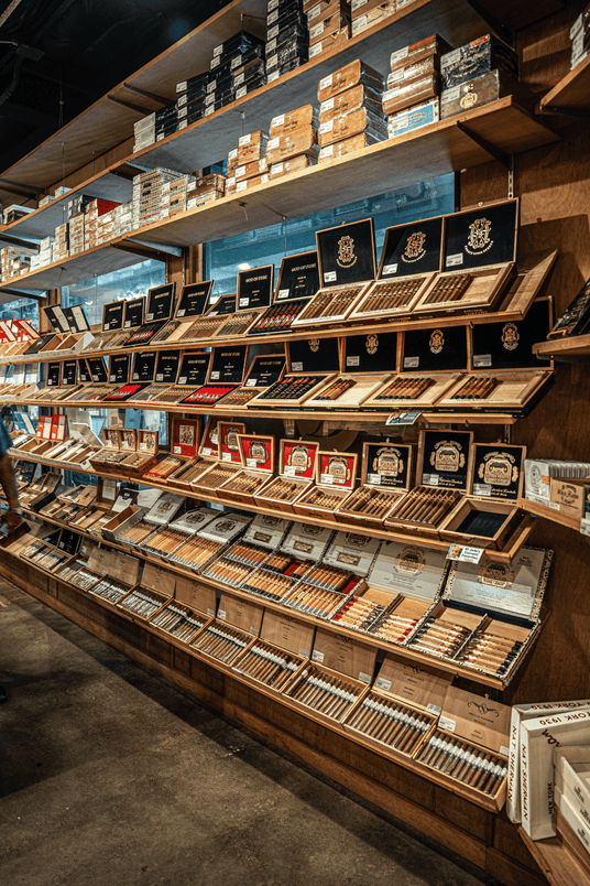 Wooden display shelves in a cigar shop filled with boxes, cartons, and open cases of cigars and cigarillos, with warm lighting and a dark store interior.